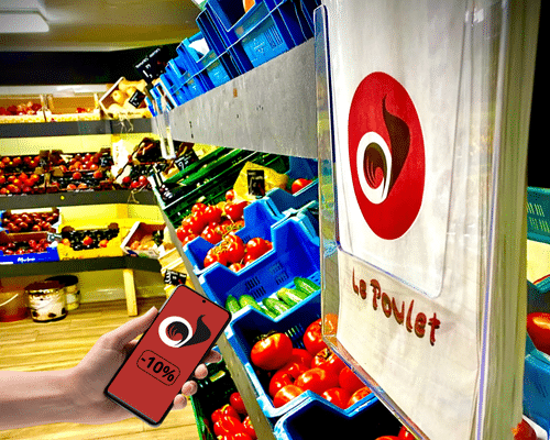 -10% Poulet Market Téléphone dans les mains qui affiche une promo de -10% sur la site du poulet market de bruxelles. Il se tient dans un rayon de légumes et fruits